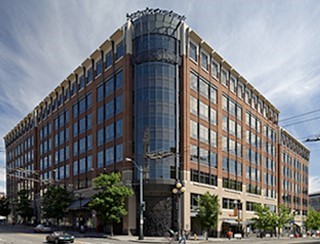 A modern multi-story building with a mix of brick and glass, featuring large windows and greenery at the base.
