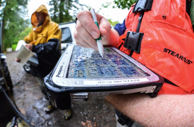 Photo captures a zoomed-in image of an electronic tablet for data collection. Only can see the persons arms holding tablet. Device is covered in water drops. Background consists of a person in a rain coat and a vehicle. 