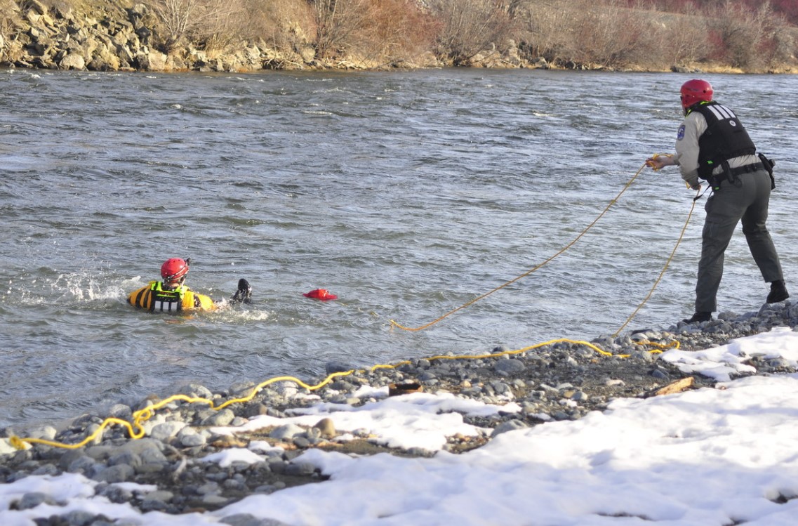 Photo of two (2) employees practicing a swift water rescue training. Employees are wearing proper personal protective equipment (PPE) near river. One employee has a long rope pulling onto an employee floating in the water.