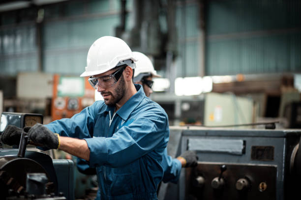 A worker in a blue jumpsuit and safety gear operates machinery in an industrial setting. Another worker is visible in the background.