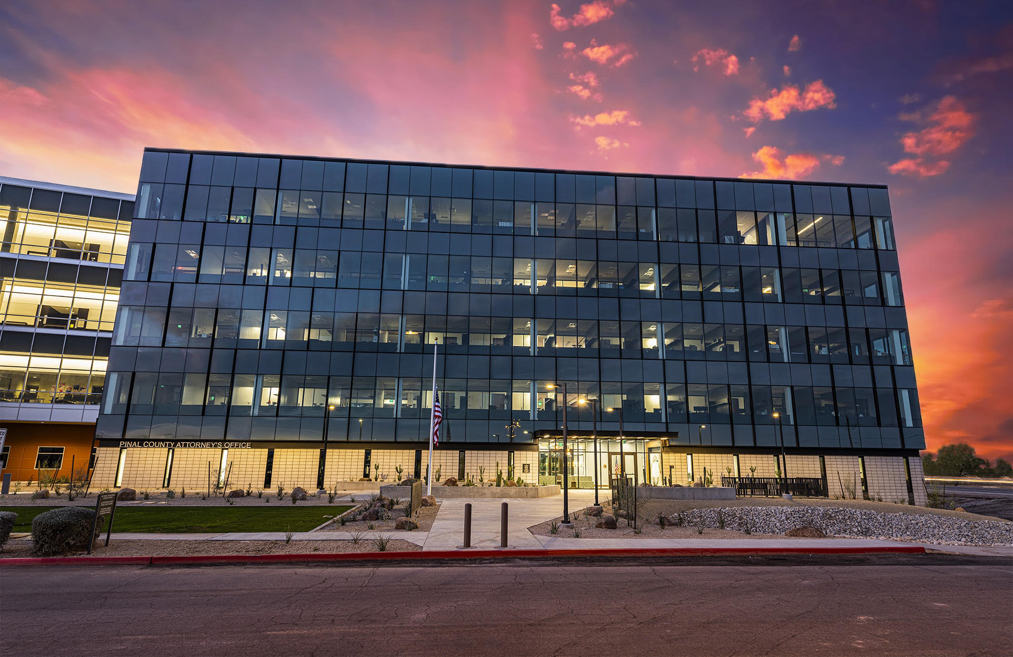 A modern glass office building with multiple floors, featuring illuminated windows and a landscaped entrance, set against a colorful sunset sky.